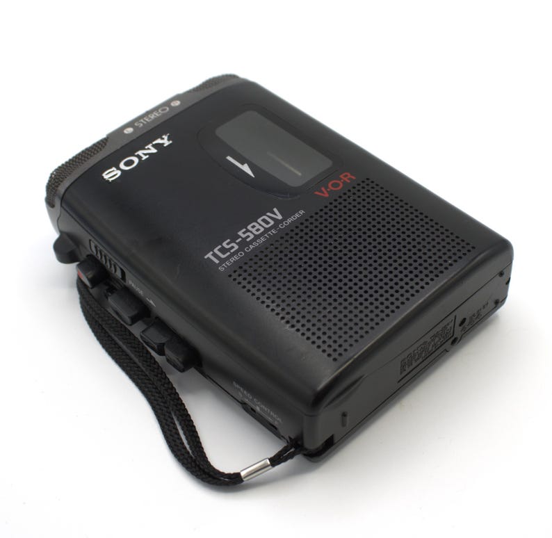 May include: Black Sony TCS-580V stereo cassette recorder with a black wrist strap. The recorder has the word "SONY" printed in white on the top left corner. The model number "TCS-580V" is printed in white on the front of the recorder.