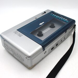 May include: Silver and blue Sony FM/AM cassette player with a black strap. The player has a clear window to view the cassette tape and a radio dial. The words "FM/AM" are printed on the front of the player.
