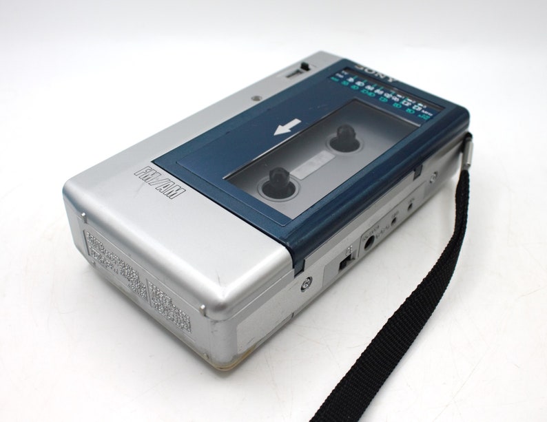 May include: Silver and blue Sony FM/AM cassette player with a black strap. The player has a clear window to view the cassette tape and a radio dial. The words "FM/AM" are printed on the front of the player.