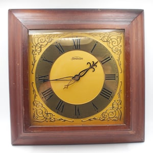 May include: A square wooden framed clock with a gold face and black Roman numeral markings. The clock face has a gold Sunbeam logo and a gold decorative border.