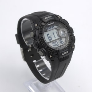 May include: Black Armitron digital watch with a black rubber strap. The watch face displays the time, date, and day of the week. Features include alarm and chronograph functions. The watch is displayed on a clear stand.
