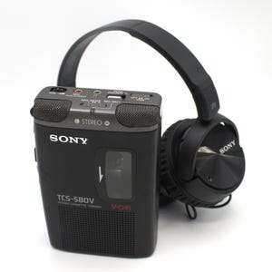 May include: A black Sony TCS-580V stereo cassette-corder with a matching black over-the-ear headphones. The cassette-corder has a built-in microphone and various control buttons. The headphones are connected to the cassette-corder.