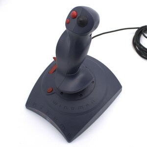 Puede incluir: Un joystick gris oscuro con botones y detalles en rojo. La base est&aacute; etiquetada como "Wingman Extreme". El joystick tiene un mango curvo y un cable negro. El dise&ntilde;o es para juegos.