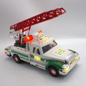 May include: A white and green toy truck with a red extending ladder. The truck has the word "HESS" in multiple places and the word "GASOLINE" on the side. The truck has working lights and red wheels.