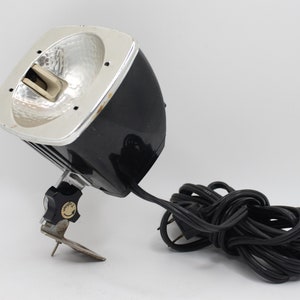 May include: A black and silver photographic spotlight with a coiled black cord. The spotlight has a swiveling arm with a knob for adjusting the angle.