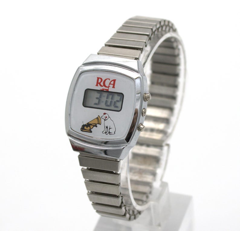 May include: A silver-tone digital wristwatch with an expandable metal link band. The white watch face features the red RCA logo, a digital time display, and a graphic illustration of a dog sitting next to a gramophone horn.