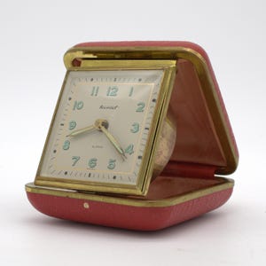 May include: A square travel alarm clock with a gold-tone metal frame and a red textured case. The clock face is off-white with light green numerals, gold hands, and the word Tourist printed in black. The clock is hinged to open from the case.