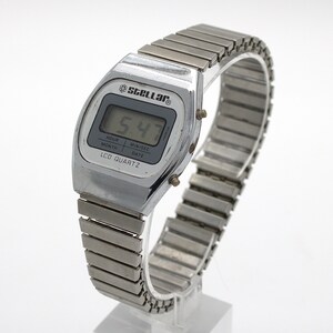 May include: A silver-tone digital wristwatch with a metallic expansion band. The watch face features the brand name Stellar, the text LCD QUARTZ, and a digital display showing 5:47. The screen includes labels for hour, month, min/sec, and date.
