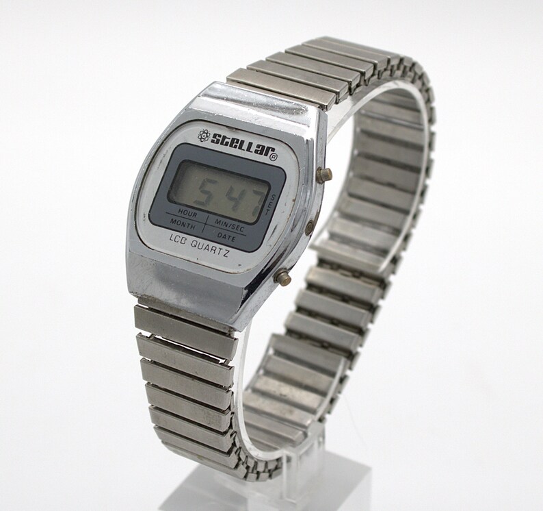 May include: A silver-tone digital wristwatch with a metallic expansion band. The watch face features the brand name Stellar, the text LCD QUARTZ, and a digital display showing 5:47. The screen includes labels for hour, month, min/sec, and date.
