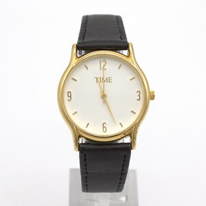 May include: A gold-toned wristwatch with a white face and black leather band. The watch face displays the word "TIME" and the numbers 3, 6, 9, and 12. The watch has a gold-colored bezel and a black leather strap.