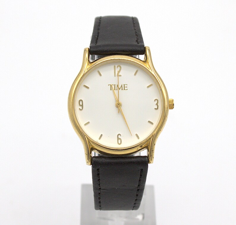May include: A gold-toned wristwatch with a white face and black leather band. The watch face displays the word "TIME" and the numbers 3, 6, 9, and 12. The watch has a gold-colored bezel and a black leather strap.