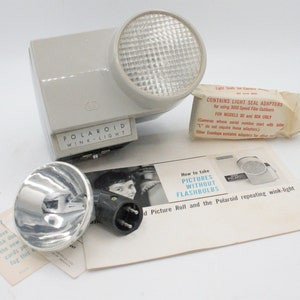 May include: A gray Polaroid Wink-Light flash unit with a silver reflector and a white instruction booklet. The booklet has the text "How to take pictures without flashbulbs" and "Polaroid repeating wink-light".