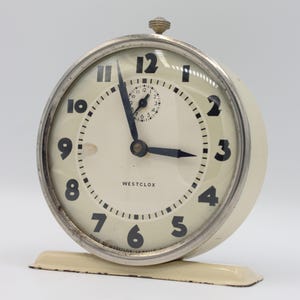 May include: A white vintage Westclox alarm clock with black numbers and hands. The clock has a silver-colored bezel and a white base.