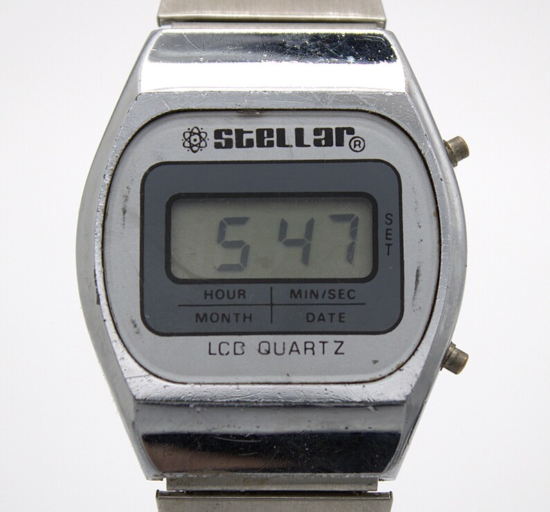 May include: A silver-toned digital wristwatch with a metallic band. The watch face features the brand name Stellar with an atom icon, a digital display showing 5:47, and the text HOUR, MIN/SEC, MONTH, DATE, and LCD QUARTZ printed on the screen.