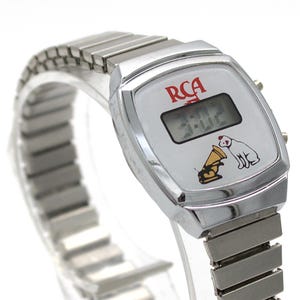 May include: A silver-tone digital wristwatch with a metallic expansion band. The white watch face features the red RCA logo, a digital time display showing 3:02, and a graphic illustration of the RCA dog listening to a gold gramophone horn.