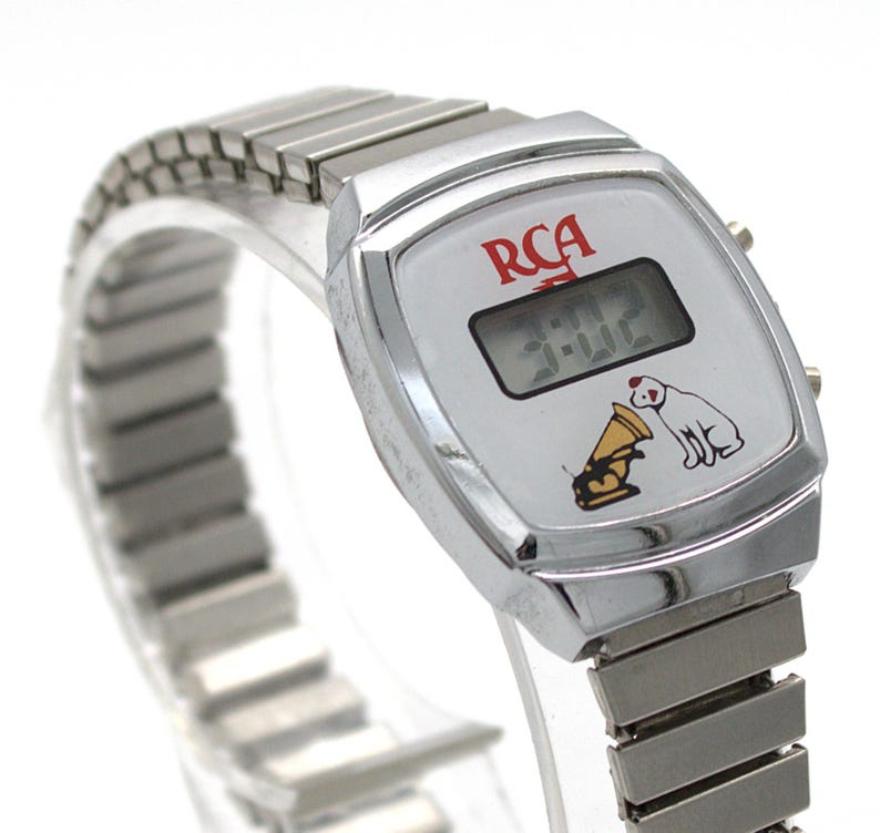 May include: A silver-tone digital wristwatch with a metallic expansion band. The white watch face features the red RCA logo, a digital time display showing 3:02, and a graphic illustration of the RCA dog listening to a gold gramophone horn.