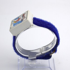 May include: Vintage children's watch with a white square face featuring a colorful illustration of a person surfing. The watch has a blue velcro strap and a clear plastic band. A retro accessory.