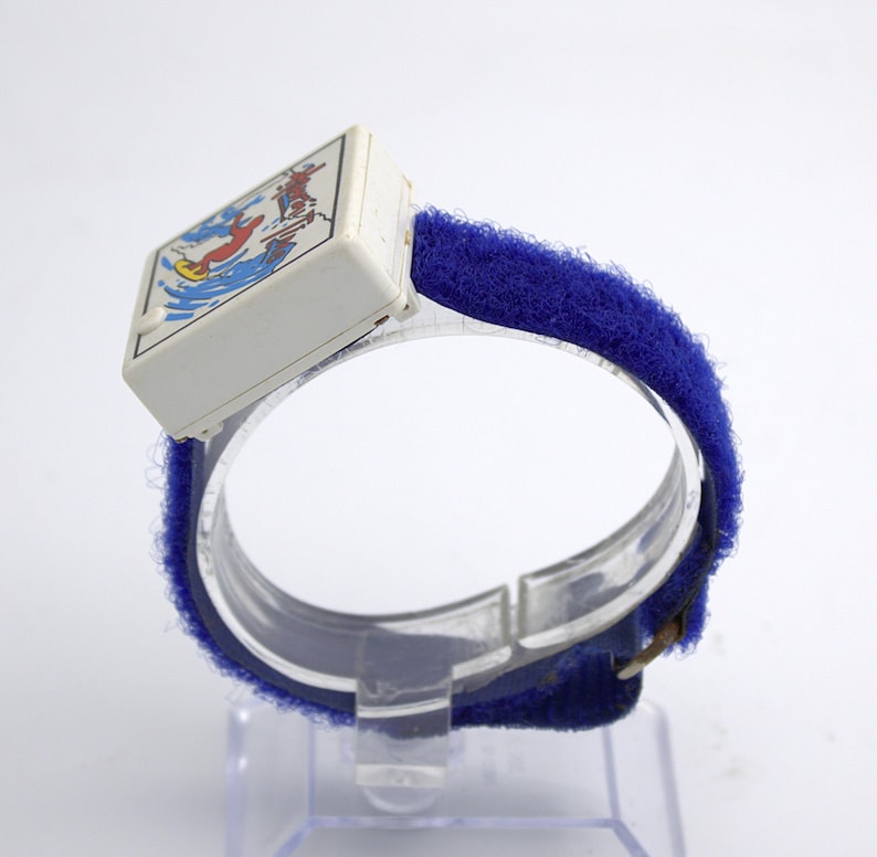 May include: Vintage children's watch with a white square face featuring a colorful illustration of a person surfing. The watch has a blue velcro strap and a clear plastic band. A retro accessory.