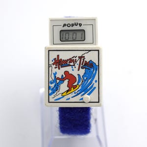 May include: A white digital watch with a blue band. The watch face displays "Hawaii Time" with a surfer graphic. The top section reads "POPUP" and shows the time 10:01.