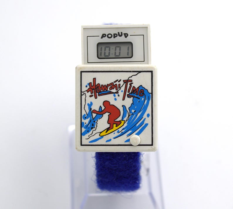May include: A white digital watch with a blue band. The watch face displays "Hawaii Time" with a surfer graphic. The top section reads "POPUP" and shows the time 10:01.