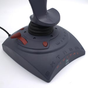 Puede incluir: Un joystick gris oscuro con una base negra y un interruptor basculante rojo. El joystick tiene un mango curvo y varios botones rojos. La base tiene el texto "Xtreme Digital" y el logotipo de Logitech.