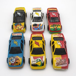 May include: A collection of six toy race cars in various colors, including yellow, red, blue, and white. Each car features a different Kellogg's cereal character and logo. The cars have black wheels and windows.
