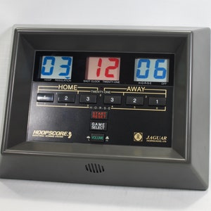 May include: A gray digital basketball scoreboard with a black display showing the score 03-12-06. The scoreboard has buttons for home and away teams, shot clock, twenty-one, H-O-R-S-E, start, reset, game select, and volume. The scoreboard is labeled "HOOPSCORE" and "JAGUAR TECHNOLOGIES LTD."