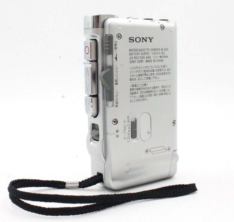 May include: A silver Sony Microcassette-Corder M-850 with a black strap. The device has a tape speed selector, play, rewind, and fast forward buttons. The text on the device reads "MICROCASSETTE-CORDER M-850 BATTERY SUPPLY 1.5Vx2 IEC, JIS R03 SIZE AAA SONY CORP MADE IN CHINA".
