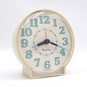 May include: Cream-colored Westclox alarm clock with a round face and a rectangular base. The clock face is off-white with teal numerals and hands. The text "Westclox" is printed on the face, and "MADE IN U.S.A." is printed at the bottom.