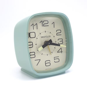 May include: A vintage, light blue Westclox alarm clock with a square face and rounded corners. The clock face is off-white with black numerals and hands. The clock reads "WESTCLOX" and "MADE IN U.S.A."