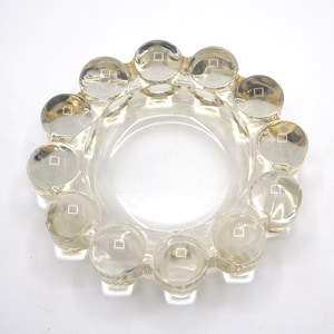May include: A clear glass ashtray with a round, scalloped edge. The ashtray has a series of small, raised circles around the rim.