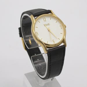 May include: A gold-toned wristwatch with a white face and black leather band. The watch face displays the word "TIME" and gold-colored numerals. The watch is displayed on a clear stand.