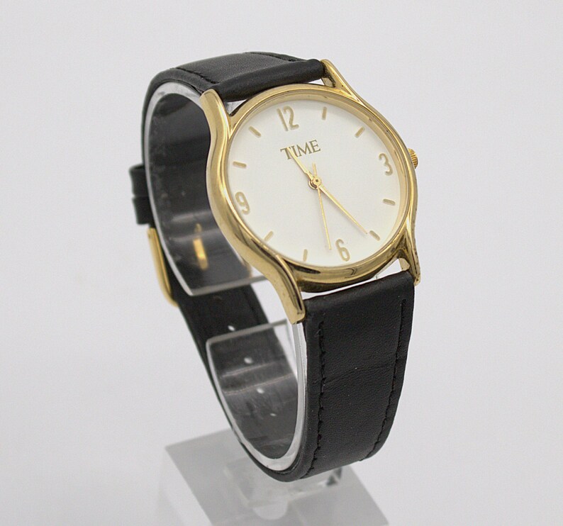 May include: A gold-toned wristwatch with a white face and black leather band. The watch face displays the word "TIME" and gold-colored numerals. The watch is displayed on a clear stand.