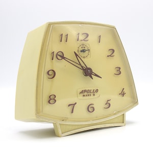 May include: A cream-colored alarm clock with a square face and a red and white "APOLLO MARK II" logo. The clock has black hands and black numbers.