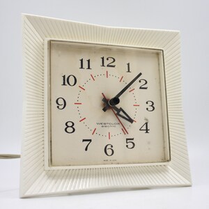 May include: A white electric clock with a square face and black hands. The clock has a white frame with a textured design. The clock face has black numbers and red markings. The clock says "Westclox Electric" and "Made in USA".