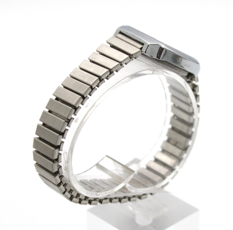 May include: A silver-toned wristwatch featuring a rectangular case and a flexible, expandable metal link bracelet. The watch has a brushed metallic finish on the band and a smooth, polished casing, presented from a side angle to show the structure.