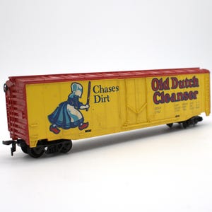 May include: A yellow and red model train boxcar featuring the Old Dutch Cleanser logo, a graphic of a person in a bonnet holding a stick, and the text Chases Dirt. The car has black wheels and couplers, with detailed side paneling and lettering.