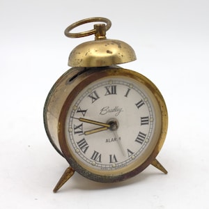 May include: A gold-toned alarm clock with Roman numeral markings on the face. The clock face reads "Bradley Alarm" and "Made in Germany".