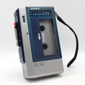 May include: A vintage Sony FM/AM cassette player, featuring a silver and dark blue design. The player has a black strap, a cassette door, and buttons on the side. The radio dial displays FM and AM frequencies.