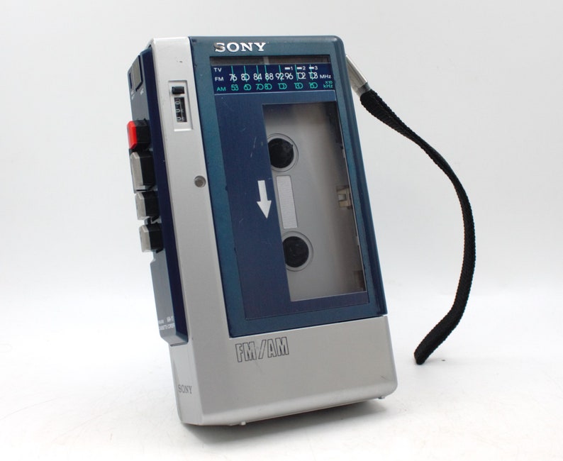 May include: A vintage Sony FM/AM cassette player, featuring a silver and dark blue design. The player has a black strap, a cassette door, and buttons on the side. The radio dial displays FM and AM frequencies.
