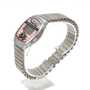 Peut inclure: Une montre num&eacute;rique argent&eacute;e avec un cadran rectangulaire. Le cadran affiche le texte "WHITE FARM EQUIPMENT" et un dessin de tracteur rouge et noir. La montre a un bracelet extensible argent&eacute;.