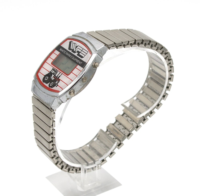 Peut inclure: Une montre num&eacute;rique argent&eacute;e avec un cadran rectangulaire. Le cadran affiche le texte "WHITE FARM EQUIPMENT" et un dessin de tracteur rouge et noir. La montre a un bracelet extensible argent&eacute;.