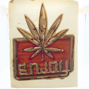 Puede incluir: Una pegatina vintage con un diseño de hoja de marihuana dorada y marrón. La palabra "Enjoy" está escrita en una fuente estilizada con un acabado metálico dorado y marrón. La pegatina está sobre un fondo beige.