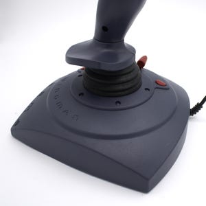 Puede incluir: Un joystick gris oscuro con botones rojos y un agarre de goma negro. La base del joystick tiene la palabra "WINGMAN" impresa. Un cable negro se extiende desde la base.