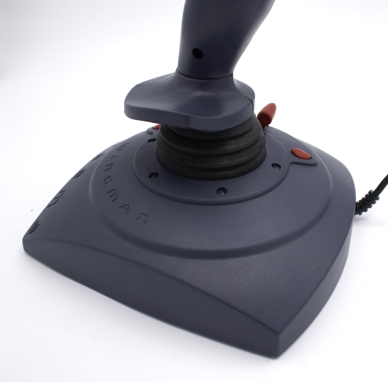 Puede incluir: Un joystick gris oscuro con botones rojos y un agarre de goma negro. La base del joystick tiene la palabra "WINGMAN" impresa. Un cable negro se extiende desde la base.