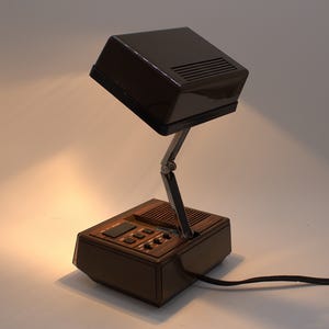 May include: A dark brown, rectangular desk lamp with a hinged, adjustable metal arm. The base features a wood-grain panel, a speaker grille, and several black control buttons. The lamp head is a solid, dark brown plastic housing with vents.