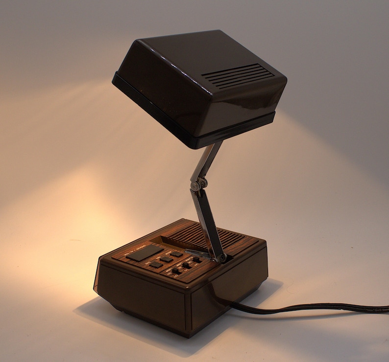 May include: A dark brown, rectangular desk lamp with a hinged, adjustable metal arm. The base features a wood-grain panel, a speaker grille, and several black control buttons. The lamp head is a solid, dark brown plastic housing with vents.