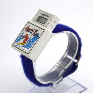 May include: A vintage white PopUp digital watch with a blue fabric band. The watch face features a surfer graphic with the words "Hang Ten Time". The digital display shows the time.