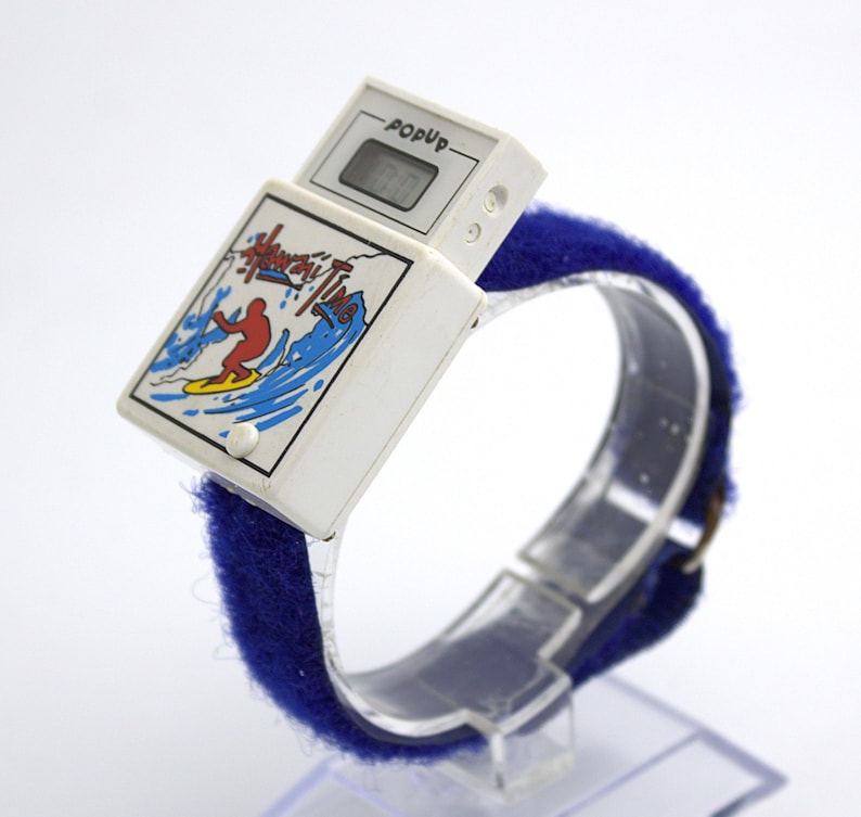 May include: A vintage white PopUp digital watch with a blue fabric band. The watch face features a surfer graphic with the words "Hang Ten Time". The digital display shows the time.