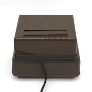 May include: A rectangular brown electronic device with a glossy finish and a black horizontal band separating the top and bottom sections. The top surface features a series of parallel ventilation slats, and a black power cord extends from the base.